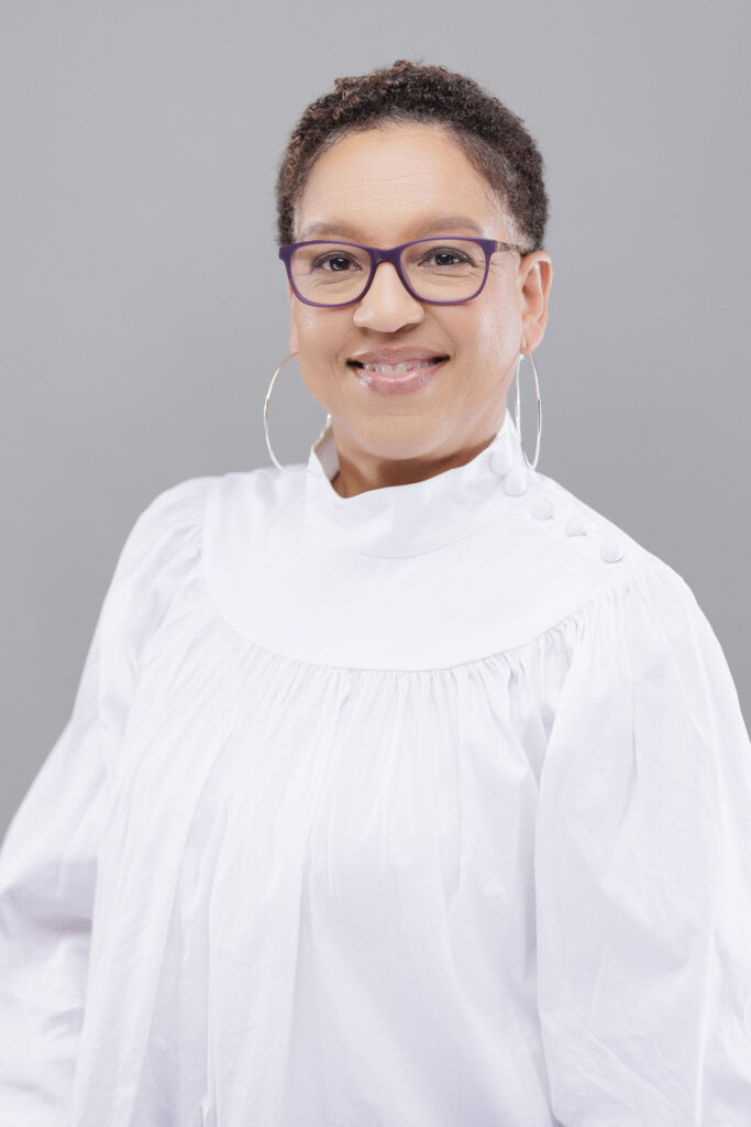 a light skinned black woman smiles at the camera she wears a white top, has short hair, purple glasses and large silver hoop earrings